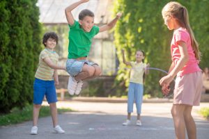 jumping enthusiastic fair haired boy bouncing up joyful friends with skipping rope park sunny day